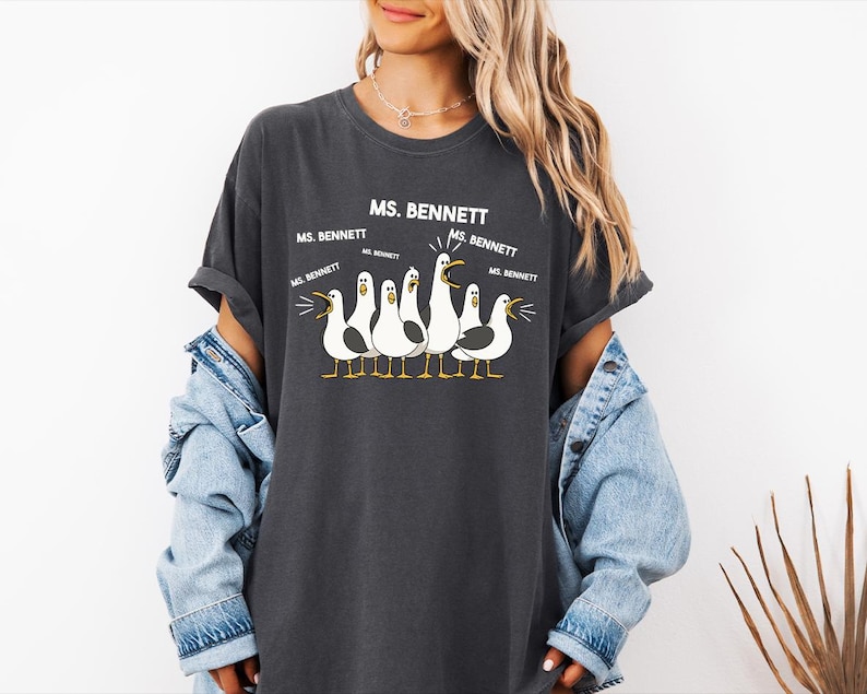 A woman wearing a gray t-shirt with a graphic of seagulls and text that says "MS. BENNETT" above them.