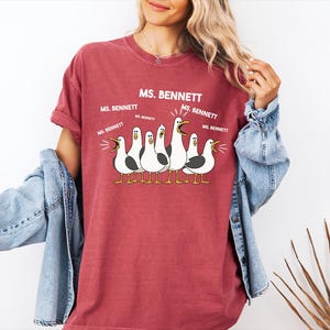A woman wearing a red t-shirt with a graphic of a group of seagulls and text that says "MS. BENNETT" and "MS. BENNETT" repeated several times.