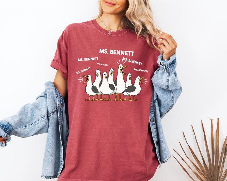 A woman wearing a red t-shirt with a graphic of a group of seagulls and text that says "MS. BENNETT" and "MS. BENNETT" repeated several times.