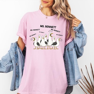 A woman wearing a pink t-shirt with a cartoon of seagulls and text that says "MS. BENNETT MS. BENNETT".