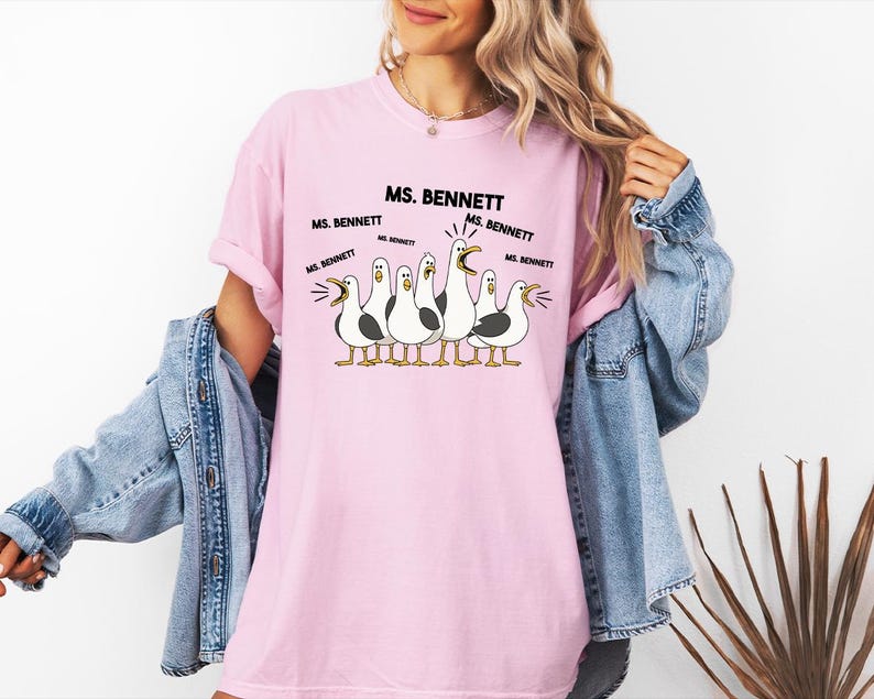 A woman wearing a pink t-shirt with a cartoon of seagulls and text that says "MS. BENNETT MS. BENNETT".