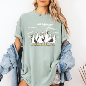 A woman wearing a green t-shirt with a graphic of a group of seagulls and text that says "MS. BENNETT MS. BENNETT".