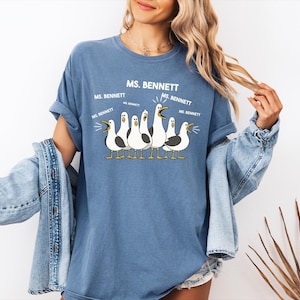 A woman wearing a blue t-shirt with a graphic of seagulls and text that says "MS. BENNETT MS. BENNETT" along with a jacket draped over her shoulders.