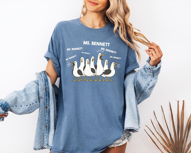 A woman wearing a blue t-shirt with a graphic of seagulls and text that says "MS. BENNETT MS. BENNETT" along with a jacket draped over her shoulders.