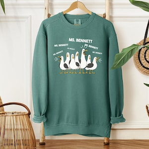 A green sweatshirt with a graphic design of five seagulls and text that says "MS. BENNETT" is hanging on a wooden hanger.