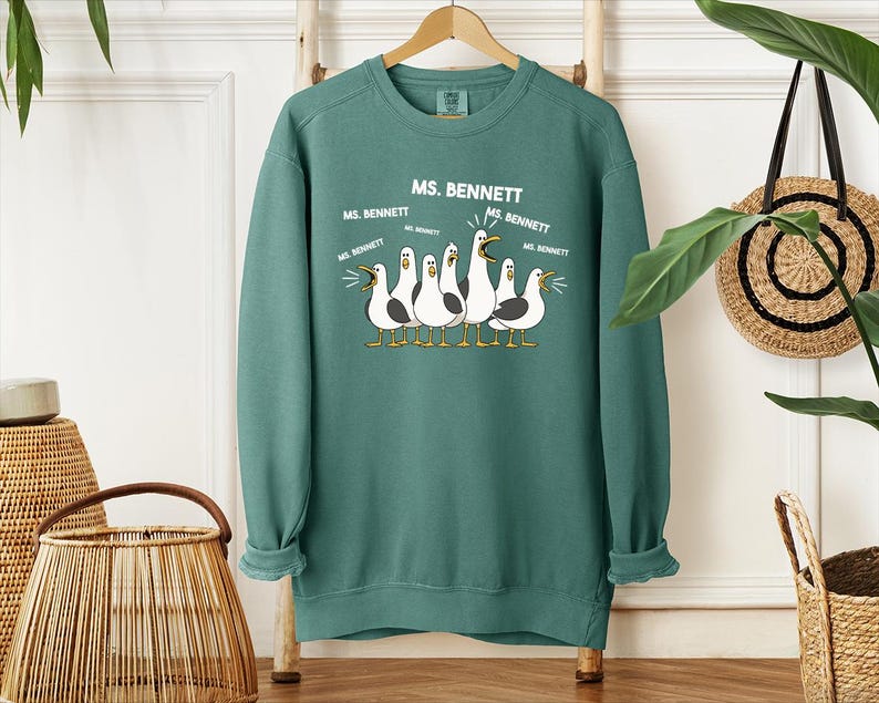 A green sweatshirt with a graphic design of five seagulls and text that says "MS. BENNETT" is hanging on a wooden hanger.