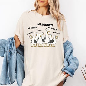 A woman wearing a white t-shirt with a graphic of a group of seagulls and text that says "MS. BENNETT MS. BENNETT".