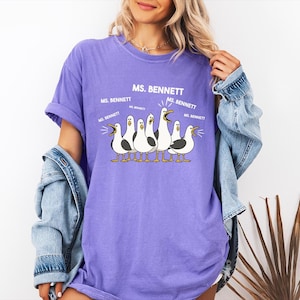 A woman wearing a purple t-shirt with a graphic of a group of seagulls and text that says "MS. BENNETT".
