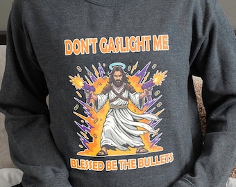 May include: Dark gray sweatshirt with the text "DON'T GASLIGHT ME" above an illustration of a figure holding two guns. The illustration is surrounded by orange and purple flames, with the text "BLESSED BE THE BULLETS" below.