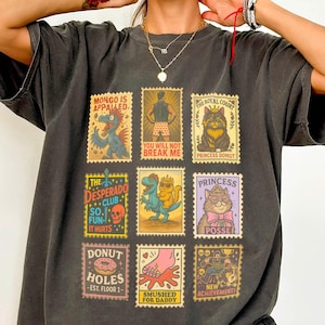 May include: Dark gray t-shirt featuring nine colorful postage stamp designs with various illustrations and text. The stamps include images of a monster, a man in swim trunks, a cat, a dinosaur, and a cat wearing glasses. Text on the stamps includes "MONGO IS APPALLED" and "DONUT HOLES".