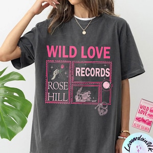May include: A dark gray t-shirt with a pink graphic print that says "Wild Love Records Rose Hill". The graphic features a hand holding a rose, a vintage record player, and a heart.