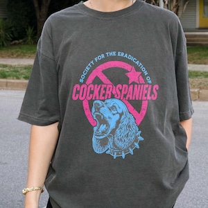 May include: Dark grey t-shirt featuring a graphic of a Cocker Spaniel with the text "SOCIETY FOR THE ERADICATION OF COCKER SPANIELS". The dog graphic is blue with a pink circle and star design.