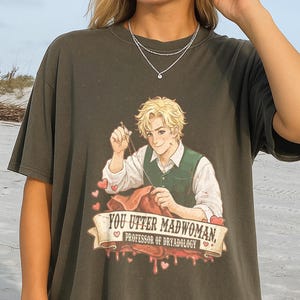 May include: Dark olive green t-shirt featuring a graphic of a blonde-haired person in a green vest, sewing. The text on the banner reads "YOU UTTER MADWOMAN, PROFESSOR OF DRYADOLOGY." Red hearts and blood-like drips are also in the design.
