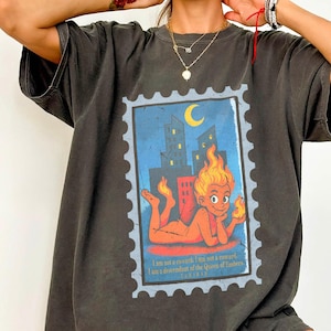May include: Dark grey t-shirt featuring a colorful stamp design. The stamp depicts a cartoon figure with flames for hair, a cityscape, and the text "I am not a reward. I am a descendant of the Queen of Unrest."