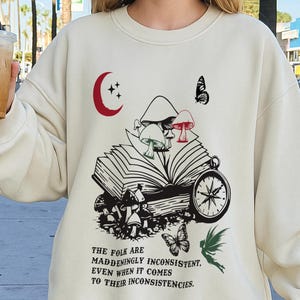 May include: Cream-colored sweatshirt with a whimsical graphic featuring mushrooms, an open book, a compass, and butterflies. The text reads, "The folk are maddeningly inconsistent, even when it comes to their inconsistencies."