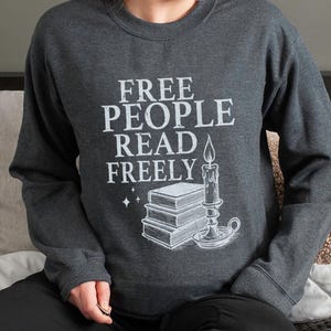 May include: Dark gray sweatshirt with the text "FREE PEOPLE READ FREELY" in white. The graphic includes a stack of books, a candle, and stars. The sweatshirt is long-sleeved and has a crew neck.