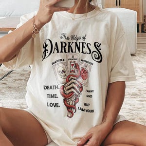 May include: Cream-colored t-shirt with the text "The Edge of Darkness" and tarot card graphics. The design includes a skull, a snake, and a heart. The shirt also features the words "Death, Time, Love" and "I am not good, but I am yours."