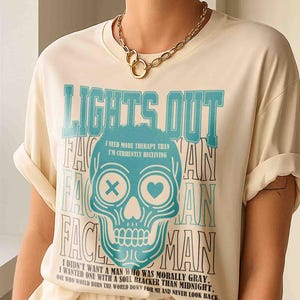 Pu&ograve; includere: T-shirt color crema con grafica a teschio turchese e le parole "LIGHTS OUT" e "VAIN". Il teschio ha una X per un occhio e un cuore per l'altro. La maglietta ha anche il testo "I need more therapy than I'm currently receiving."