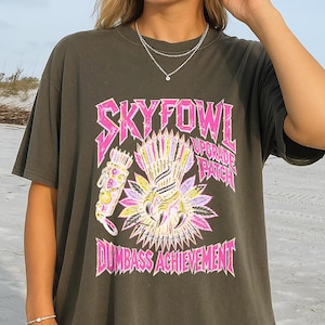 May include: A dark charcoal gray short-sleeve t-shirt featuring a graphic print in pink, yellow, and white. The text reads SKYFOWL UPGRADE PATCH and DUMBASS ACHIEVEMENT, accompanied by an illustration of a bird claw and a quiver of arrows.