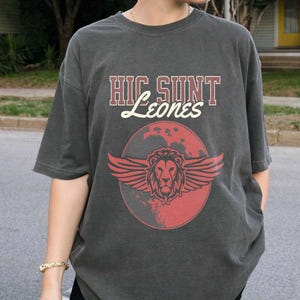 May include: A charcoal gray short-sleeve t-shirt featuring the text HIC SUNT Leones in a mix of block and script fonts. Below the text is a graphic of a winged lion head centered over a red circular shape that resembles a stylized planet.