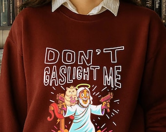 May include: Maroon sweatshirt with the text "DON'T GASLIGHT ME" above a cartoon illustration of a figure holding two guns and a cat. The figure has a halo and is wearing a white robe. The sweatshirt has long sleeves.