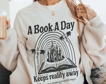 May include: A white sweatshirt with a black graphic print of a castle, a rainbow, and a book. The text on the graphic reads "A Book A Day Keeps Reality Away" and "Books are a uniquely portable magic."