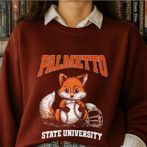 May include: A maroon sweatshirt with the text "PALMETTO STATE UNIVERSITY" in orange lettering. A cartoon fox holding a baseball is centered below the text. The sweatshirt has long sleeves and a white collared shirt underneath.