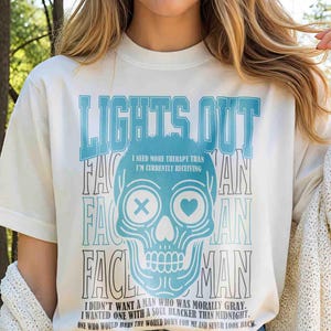 Pu&ograve; includere: T-shirt color crema con grafica di teschio blu e la scritta "LIGHTS OUT". La grafica include il testo "I NEED MORE THERAPY THAN I'M CURRENTLY RECEIVING" e una citazione sul desiderio di un'anima pi&ugrave; oscura.