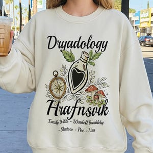 May include: Cream-colored sweatshirt with the words "Dryadology" and "Hrafnsvik" in black script. The design includes a compass, a potion bottle, and mushrooms. The names Emily Wilde, Wendell Bumbleby, Shadow, Poe, and Lisa are also listed.