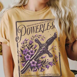 May include: Mustard yellow t-shirt featuring a graphic design with the word "POWERLESS" in a vintage style. The design includes a sword, purple flowers, and text that reads "Strength is not in what you can do, but in what you choose to fight."