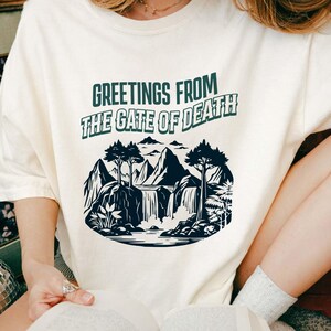 May include: Cream-colored t-shirt with the text "GREETINGS FROM THE GATE OF DEATH" in green. The shirt features a black and white graphic of a mountain landscape with waterfalls and trees. The person is holding a book.