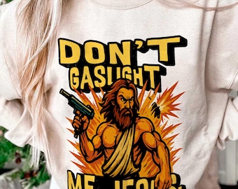 May include: Beige sweatshirt with the text "DON'T GASLIGHT ME, JESUS" in yellow and black. The graphic features a muscular Jesus figure holding a gun, with an orange explosion background.
