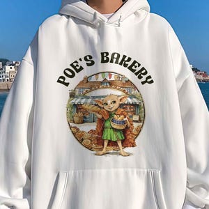 May include: White hoodie with a front pocket and a graphic that reads "Poe's Bakery." The graphic features a cartoon character in a green outfit holding a basket of baked goods in front of a bakery. The hoodie has white drawstrings and white cuffs.