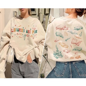 May include: A cream-colored sweatshirt with the text "LIBRARIES ARE FOR everyone" in colorful letters and stars. The back of the sweatshirt has a pattern of books, pens, and feathers.