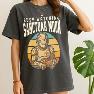 May include: Dark gray t-shirt with the text "BUSY WATCHING SANCTUAR MOON" above a retro-style graphic of a robot holding a game controller. The robot is set against a circular background with orange, yellow, and blue stripes.