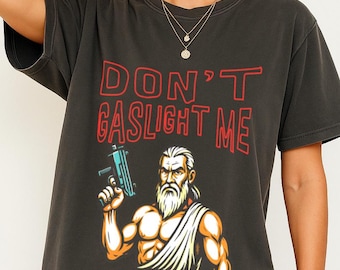 May include: Dark gray t-shirt with the text "DON'T GASLIGHT ME" in red. The shirt features a graphic of a muscular man with a white beard holding a blue gun. The man is wearing a white toga.