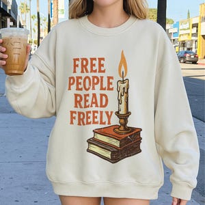 May include: A cream-coloured sweatshirt featuring the orange text "FREE PEOPLE READ FREELY". A candle with a lit flame is placed on a stack of books. The sweatshirt is a casual, comfortable style.