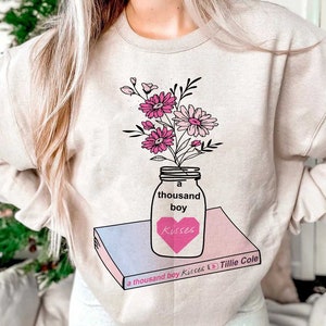 May include: A beige sweatshirt with a graphic design of a mason jar filled with pink and white flowers. The jar is labeled "a thousand boy Kisses" and is sitting on top of a book with the title "a thousand boy Kisses by Tillie Cole".