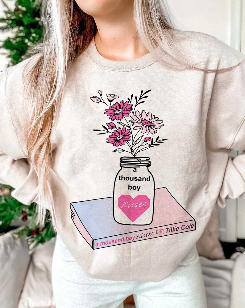 May include: A beige sweatshirt with a graphic design of a mason jar filled with pink and white flowers. The jar is labeled "a thousand boy Kisses" and is sitting on top of a book with the title "a thousand boy Kisses by Tillie Cole".