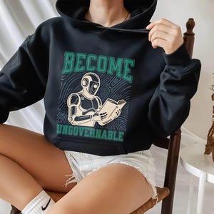 May include: Black hoodie with the text "BECOME UNGOVERNABLE" in green. The design shows a robot reading a book. The sweatshirt is dark and has a drawstring hood. The person is wearing white shorts.