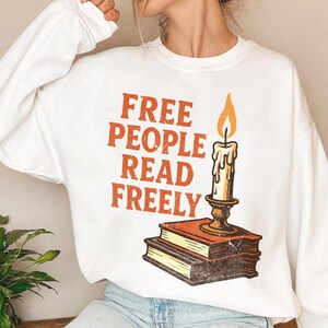 May include: White sweatshirt featuring the slogan "FREE PEOPLE READ FREELY" in orange. A candle with an orange flame is placed on two books. The design has a vintage, distressed appearance.