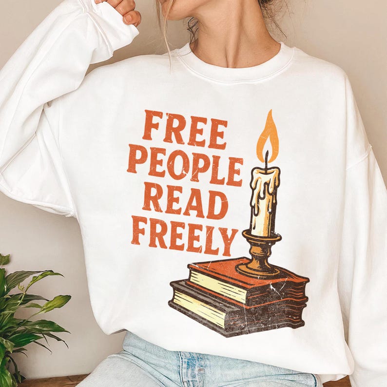 May include: White sweatshirt featuring the slogan "FREE PEOPLE READ FREELY" in orange. A candle with an orange flame is placed on two books. The design has a vintage, distressed appearance.