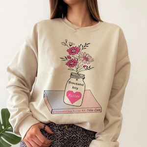 May include: A beige sweatshirt with a graphic print of a mason jar filled with pink and white flowers. The jar has the text "a thousand boy kisses" on it. The text "a thousand boy kisses Tillie Cole" is printed on a book below the jar.