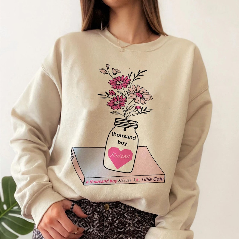 May include: A beige sweatshirt with a graphic print of a mason jar filled with pink and white flowers. The jar has the text "a thousand boy kisses" on it. The text "a thousand boy kisses Tillie Cole" is printed on a book below the jar.
