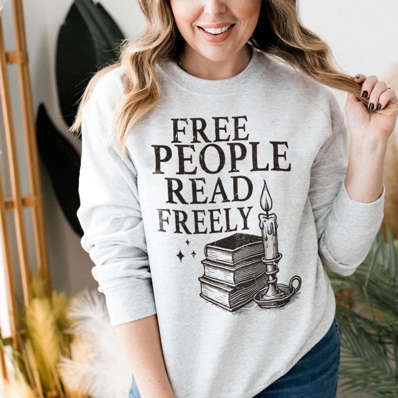 May include: A light gray sweatshirt with the text "FREE PEOPLE READ FREELY" in black. Below the text is a graphic of three books, a candle, and stars. The sweatshirt is a crew neck style.