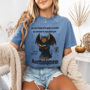 May include: A blue t-shirt featuring a graphic of a cat-like creature with bat wings, reading a book. The text on the shirt reads "You don't have to be good, or useful, for someone to care about you." and "Bortholomew."