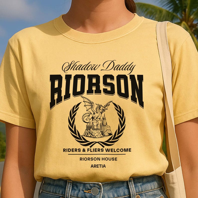 May include: Mustard yellow t-shirt with black text reading "Shadow Daddy RIORSON", above a circular emblem with a dragon and castle design, encircled by a laurel wreath. Below, it says "RIDERS & FLIERS WELCOME, RIORSON HOUSE, ARETIA."