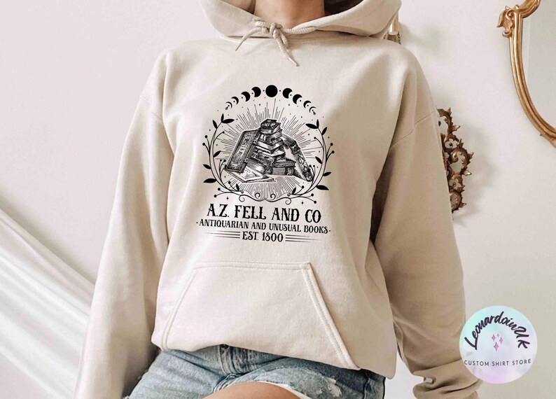 Pu&ograve; includere: Una felpa con cappuccio beige con un design grafico nero che presenta una pila di libri, una luna crescente e il testo "A.Z. Fell and Co. - Antiquarian and Unusual Books - Est 1800".