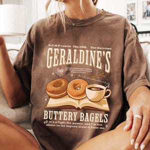 May include: Brown t-shirt featuring a graphic design with the text "GERALDINE'S BUTTERY BAGELS". The design includes illustrations of two donuts, a cup of coffee, and an open book. The shirt has a vintage, worn look.