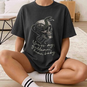 May include: Dark gray t-shirt featuring a skull with a compass design and a green snake. The shirt has the text "You're my goddamn, home, baby" in a script font. The shirt is worn by a person sitting on the floor.